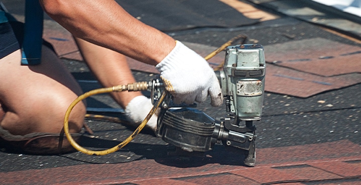 A roofer with gloves on is installing brown asphalt shingles with an air nailer finding a good roofing contractor is tricky