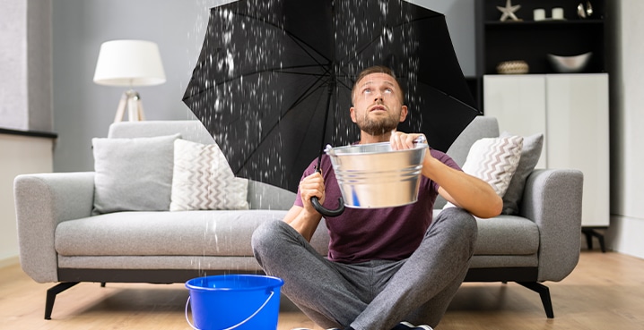 Man sitting in LR with umbrella and buckets of water roof repair flashing